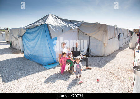 Two men sit and relax in the morning sunshine with three small children outside the rows of homes at the Ritsona Refugee Camp. Stock Photo