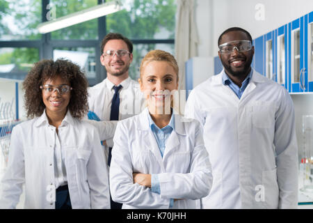 Smiling Group Of Scientists In Modern Laboratory With Female Leader, Mix Race Team Of Scientific Researchers In Lab Wearing White Coats And Protective Stock Photo