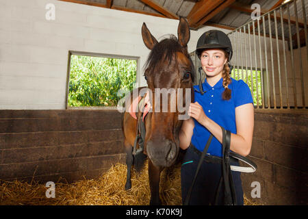 Beautiful female jockey standing inside the stable and hugging her bay horse Stock Photo