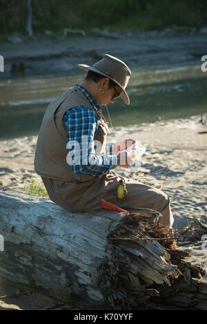 Side view of man looking at fishing bait while sitting on fallen tree trunk during sunny day Stock Photo
