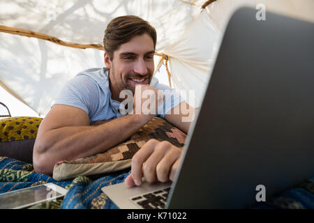 Smiling young man using laptop while lying down in tent Stock Photo