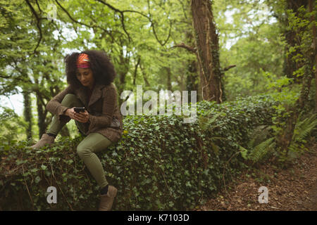 Young woman using phone while sitting on wall covered by creeper plants at forest Stock Photo