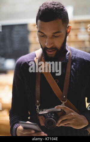 Close up of young man with camera using mobile phone while standing in cafe Stock Photo