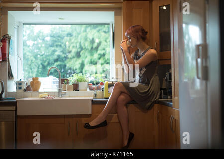 Young woman drinking coffee while sitting on counter in kitchen Stock Photo