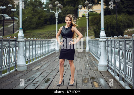 Outdoor lifestyle portrait of blonde young woman in stylish black dress staying on bridge on the street. Autumn, rainy day. Kyiv Stock Photo