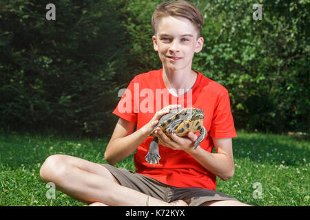 Cute teenager boy wearing red t-shirt sitting on a lawn in a summer garden holding turtle looking at camera smiling Stock Photo