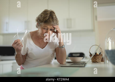Tense senior woman standing in kitchen with laptop on worktop at home Stock Photo