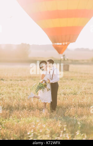 Happy newlywed couple is having fun in the sunny field. The groom is kissing the vintage dressed bride in the cheek at the background of the airballoon. Stock Photo