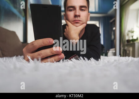 a young man looks at his smartphone at home. Man relaxing during looking at the screen while lying on sofa. Read electronic books on your phone. Stock Photo