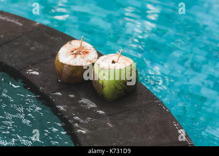Two young fresh coconuts on edge of swimming pool with crystal clean emerald water. Tropical and summer mood Stock Photo