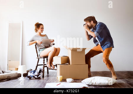 Young married couple moving in new house, woman with tablet sitting on chair, man taking pictures of her. Stock Photo