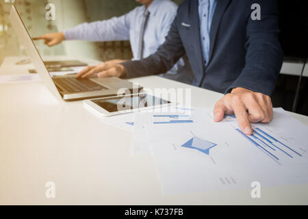 business documents on office table with laptop and digital tablet and stylus and two colleagues discussing data in the background. Stock Photo