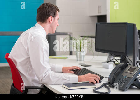 A young male office worker at his desk working in a modern office,on a computer with a blank screen Stock Photo