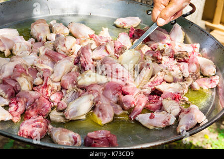 Man's hand holding a turner, tossing rabbit and chicken fresh meat slices frying in large flat paella pan. Process of cooking. Outdoors, summer day, w Stock Photo