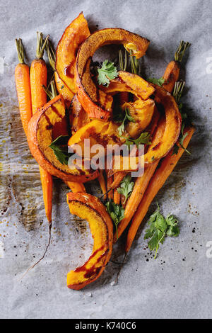 Roasted young whole carrot and sliced pumpkin with greens and sea salt on white baking paper as background. Top view with space Stock Photo