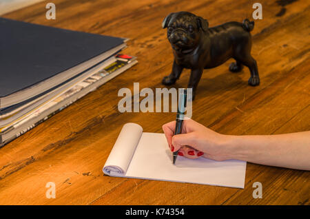 Woman hand writing on blank notepad on top of rustic wooden table with decorative objects, pug sculpture. Stock Photo