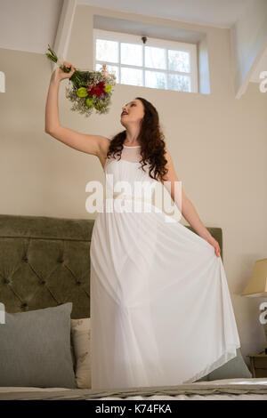 Low angle view of beautiful bride holding bouquet singing while standing on bed at home Stock Photo