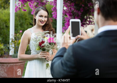 Rear view of bridegroom photographing bride holding bouquet at park Stock Photo