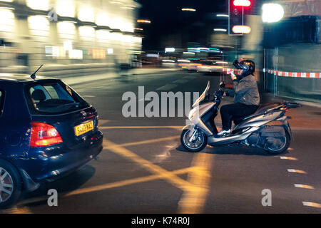 A late-night moped rider drives through Central London traffic near King's Cross station while using an app on his phone. Stock Photo