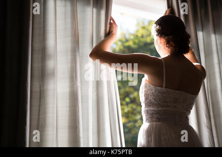 Rear view of bride in wedding dress looking through window while standing at home Stock Photo