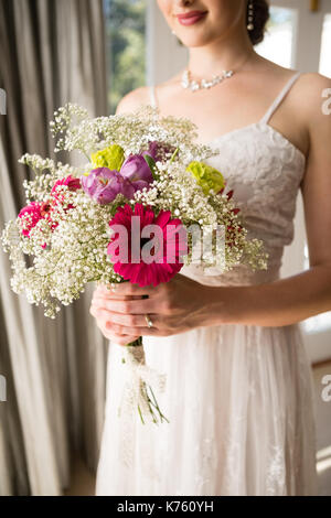 Midsection of smiling bride in wedding dress holding bouquet while standing at home Stock Photo