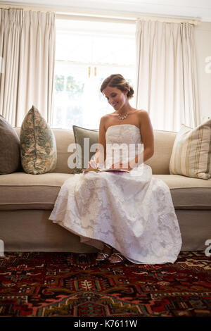 Beautiful bride writing in diary while sitting on sofa at home Stock Photo