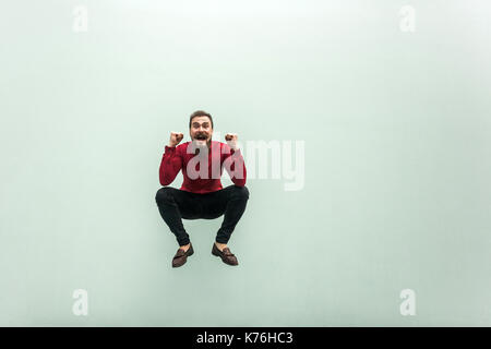 Like a frog. Jumping higher. Bearded man in red shirt celebrating hes victory, looking at camera and toothy smile. Studio shot, isolated on gray backg Stock Photo