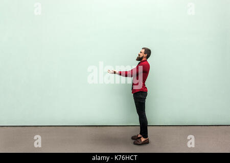 Young adult bearded man holding something heavy and showing it. Outdoor, gray wall Stock Photo