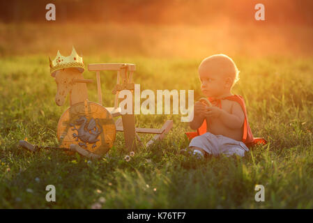 Little boy king sits with a horse toys outdoors at sunset with the light beam. Prince eats resting on the grass. Stock Photo