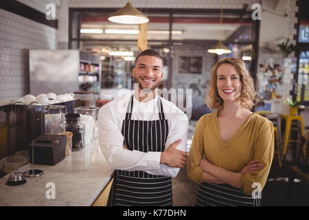 Portrait of smiling young wait staff standing with arms crossed by counter in coffee shop Stock Photo