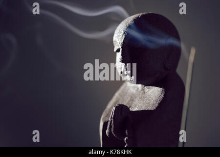 Praying Buddha statue with burning incense smoke in dramatic dim light on dark gray background Stock Photo