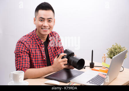 Professional photographer. Portrait of confident young man in shirt holding hand on camera while sitting at his desk. Stock Photo