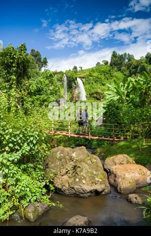 A couple standing on a wooden bridge over a river viewing Sipi waterfalls in the background, Kenya Stock Photo