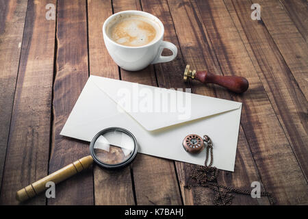 Vintage still life with postal accessories. Blank envelope, magnifier, stamp, clock and coffee cup on wooden table background.