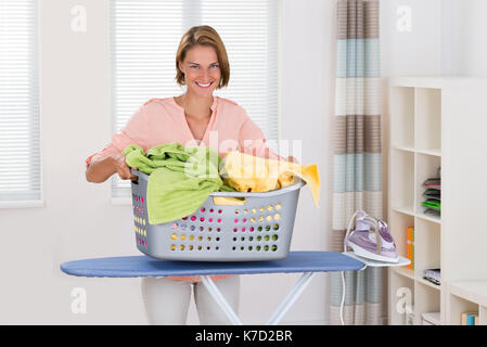 Young Happy Woman With Clothes Basket And Electric Iron On Ironing Board Stock Photo