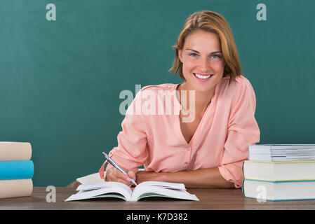 Young Happy Female Teacher Studying At Desk In Classroom Stock Photo
