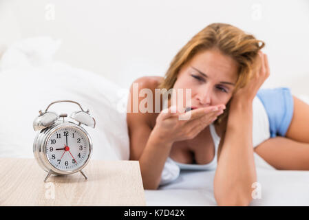 Young Woman Yawning With Classic Alarm Clock On Nightstand Stock Photo