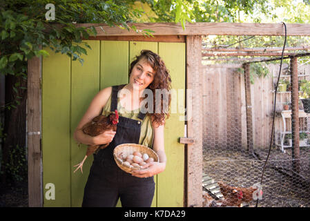 Portrait of happy woman holding hen and egg basket while standing against birdcage at farm Stock Photo
