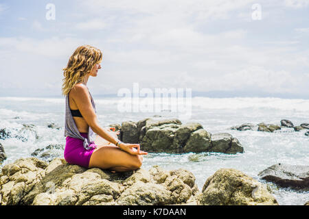 Side view of mid adult woman meditating in lotus position on rocks at beach Stock Photo