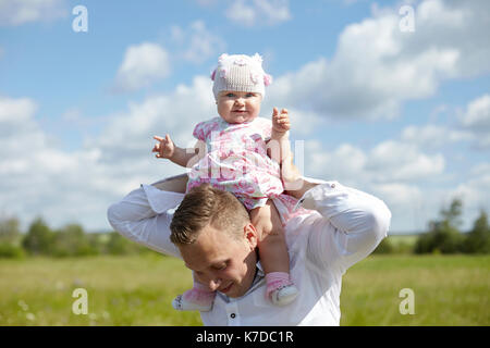 Portrait of cute baby girl sitting on father's shoulder at field against sky during sunny day Stock Photo