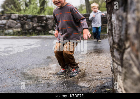 Midsection of boy jumping in puddle with brother in background Stock Photo