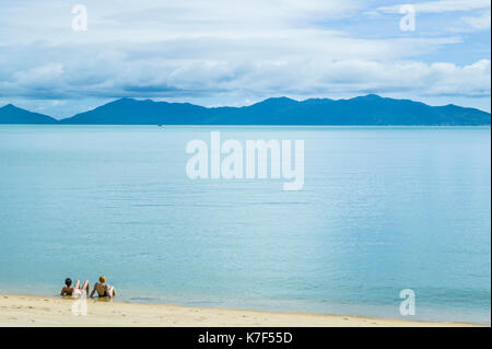 A couple relaxing in the shallow water just off a beach in Koh Samui, Thailand. Koh Phangan is the island in the distance. Stock Photo