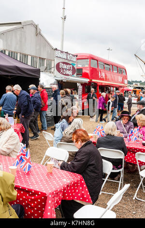 England, Chatham Dockyard. Event, Salute to the 1940s. People, many in 1940s fashion, sitting and standing around tables drinking teas. Red London bus café in background. Stock Photo