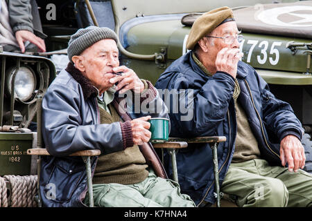 England, Chatham dockyard. Two old men in their sixties. wearing US world war two army clothing sitting with mug of tea and smoking while watching the world go by. Stock Photo