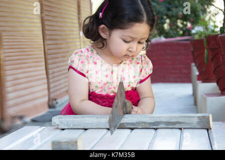 Little girl carpenter sawing wood plank with a handsaw outdoors. Indian, Asian. Stock Photo