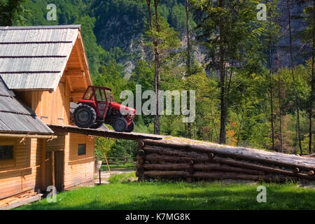 Rustic old red tractor on ramp of wooden barn, woods and mountains in background Stock Photo