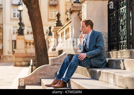 A middle age businessman siting on the stairs with a laptop and a phone in his hand and thinking Stock Photo