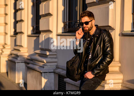 A handsome young hipster man leaning to the wall of a house on a sunny street while talking on his smartphone Stock Photo
