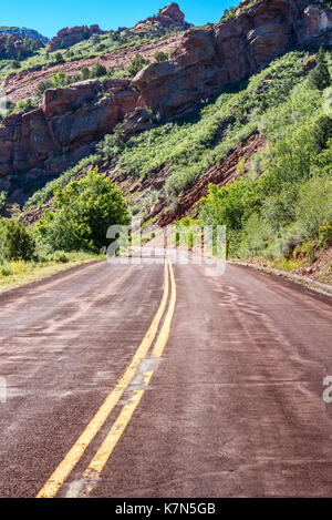 Paved mountain road red asphalt double yellow lines red sandstone mountain green trees blue sky. Stock Photo