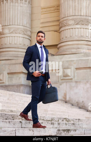 A handsome young businessman walking down on the stairs while holding his phone and his bag Stock Photo